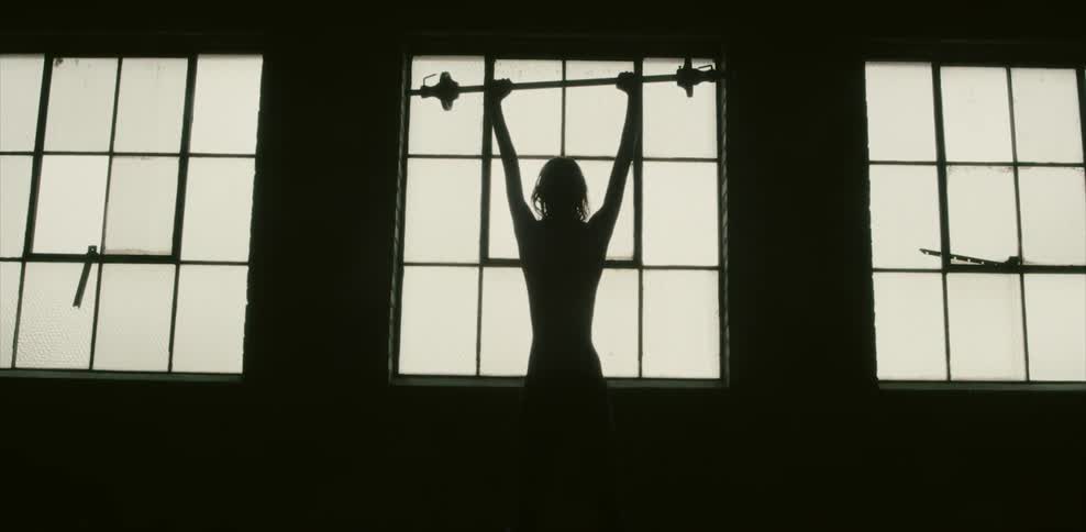 A silhouetted man lifts a barbell overhead in front of a window