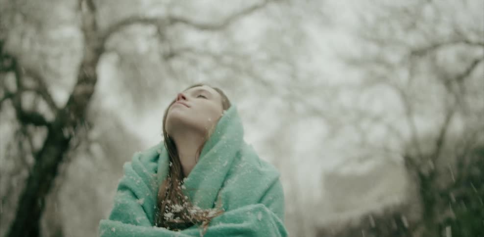 Woman covered with a blanket stands outside looking up at falling snow