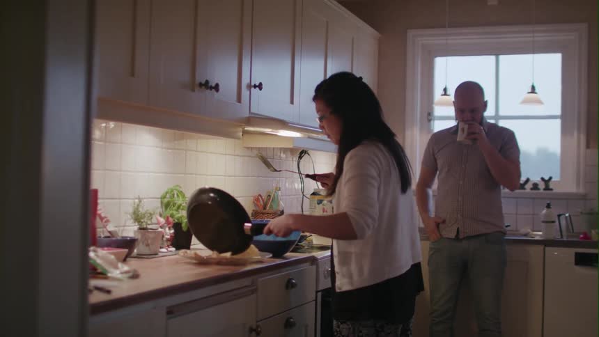 A woman fixes a meal in the kitchen as a man sips from a mug