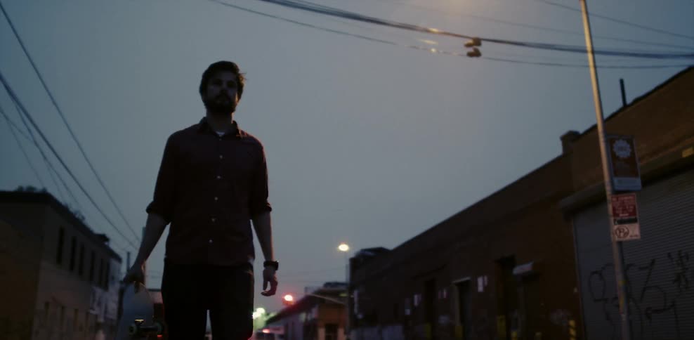 A man walking in a street with warehouses gets on his skateboard