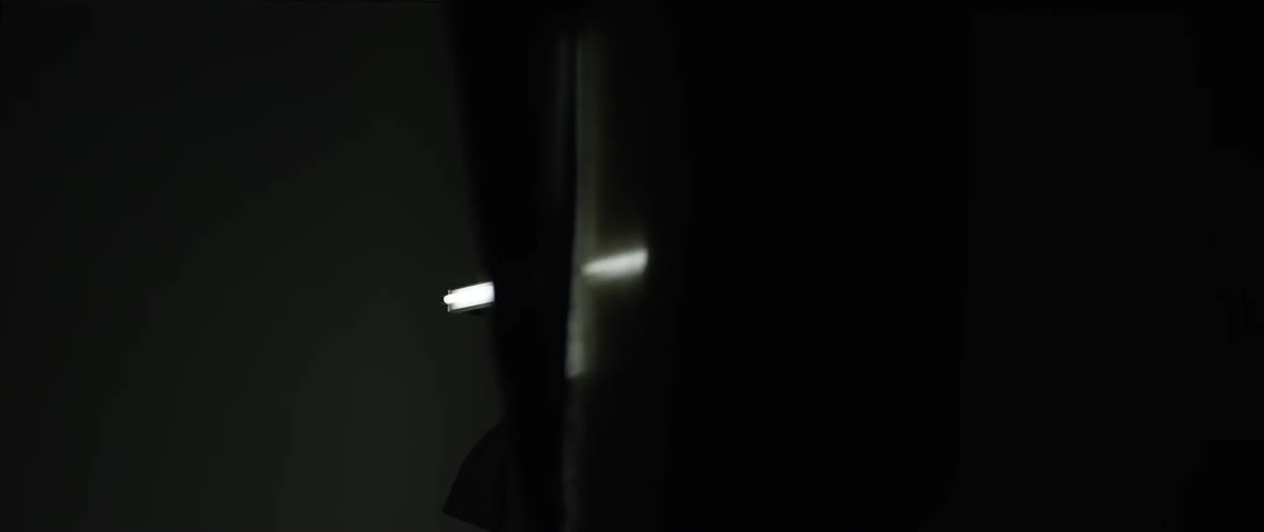 A white cross is seen illuminated against a black background with thin worn curtains blowing in front of it