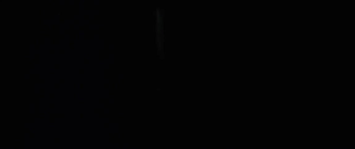 Thin, worn curtains obscure the view of a white illuminated cross against a black background