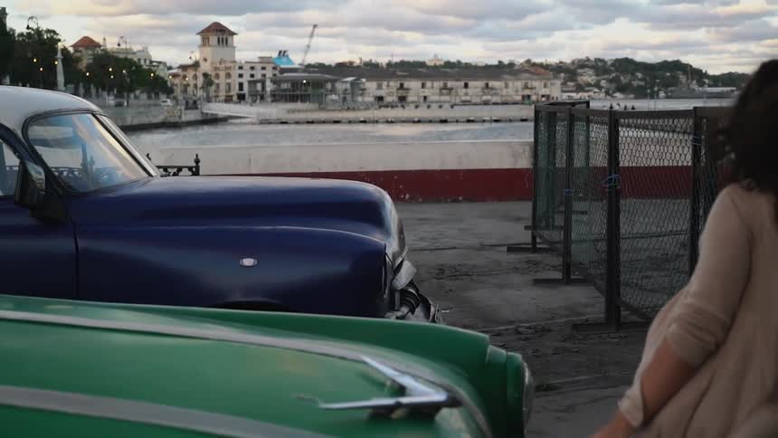 Woman touches a classic car next to a body of water and a city
