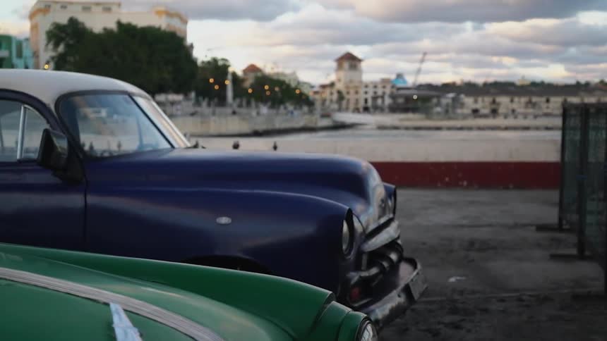 Two classic cars parked at the edge of a body of water with a city behind