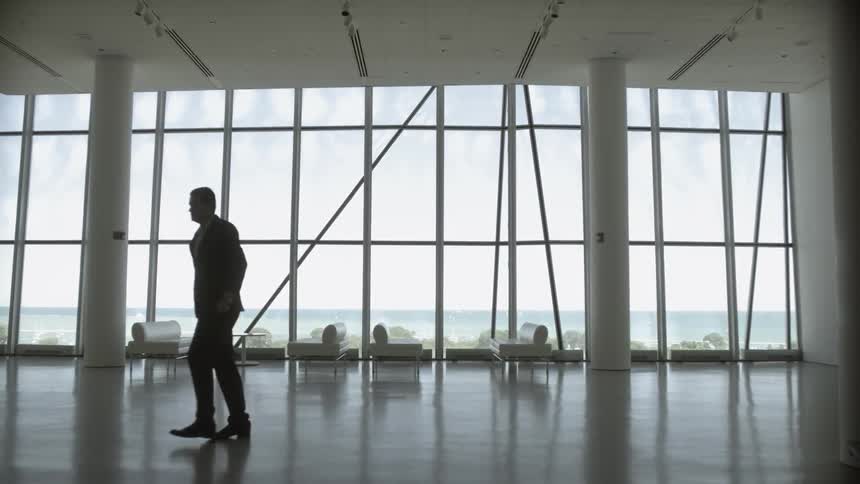 A man in a suit stands in a windowed lobby, checks his watch and then walks away