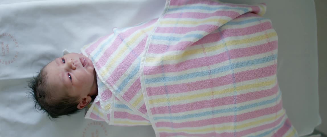 A swaddled newborn baby lies in its hospital crib turning its head