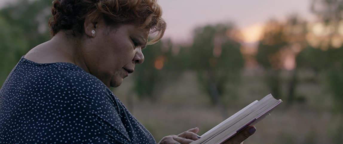 An hispanic woman is reading out loud from a book outside at dusk