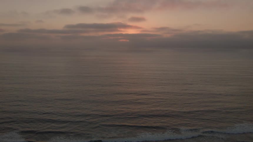 A view of the sun setting on a cloudy evening as waves crash and foam