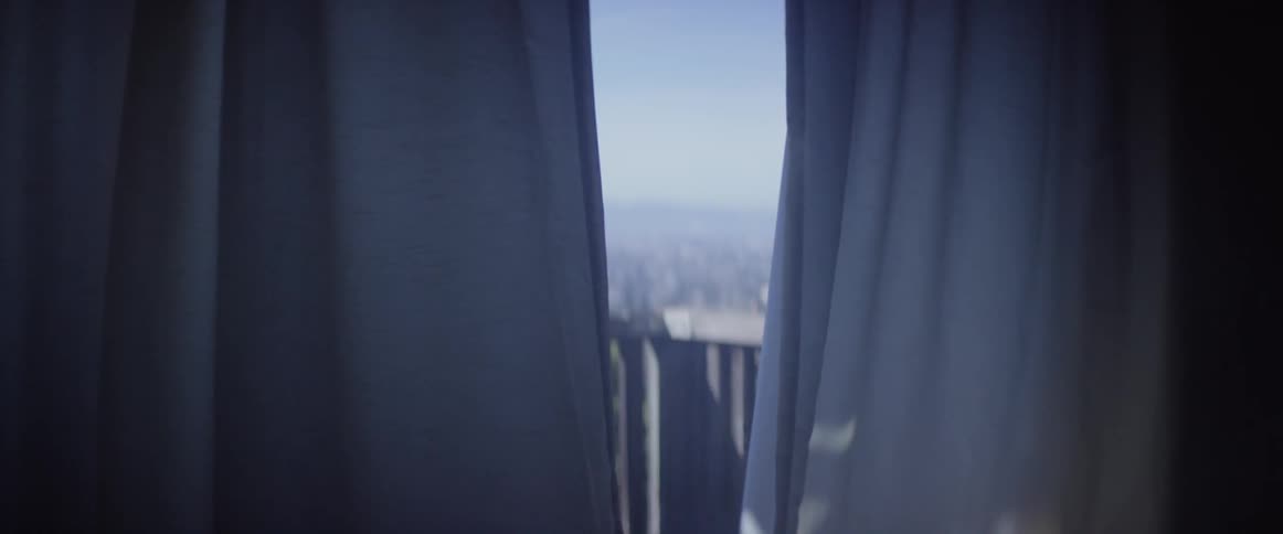 Curtains blow in the breeze and part to reveal a balcony looking out on an old city