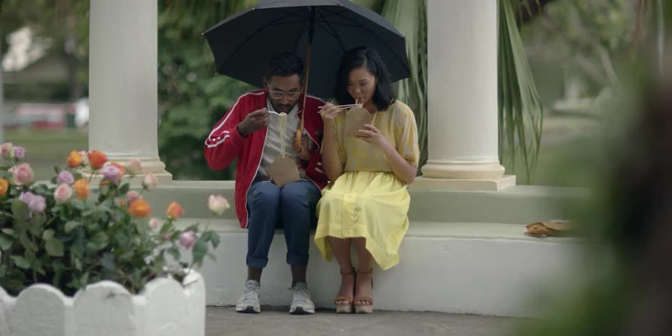 A man and woman sit beneath an umbrella while they eat takeout with chopsticks