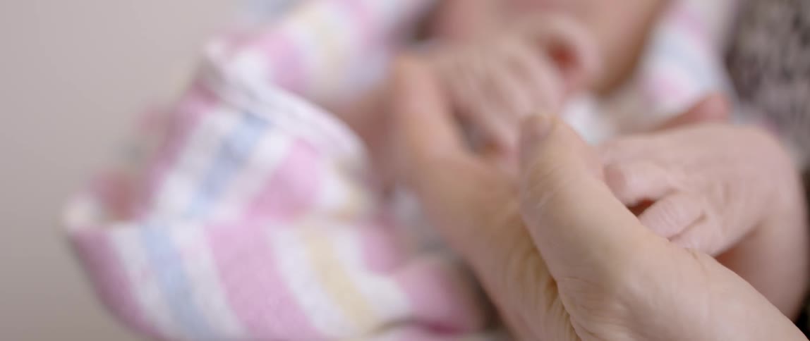 An adult holds the hand of a newborn wrapped in a blanket