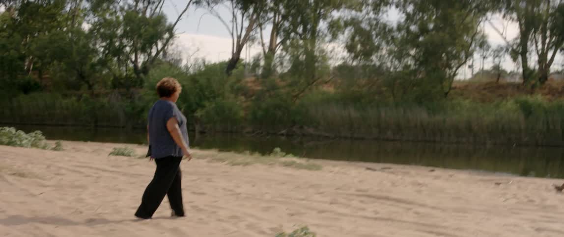 A woman walks across sand toward a river