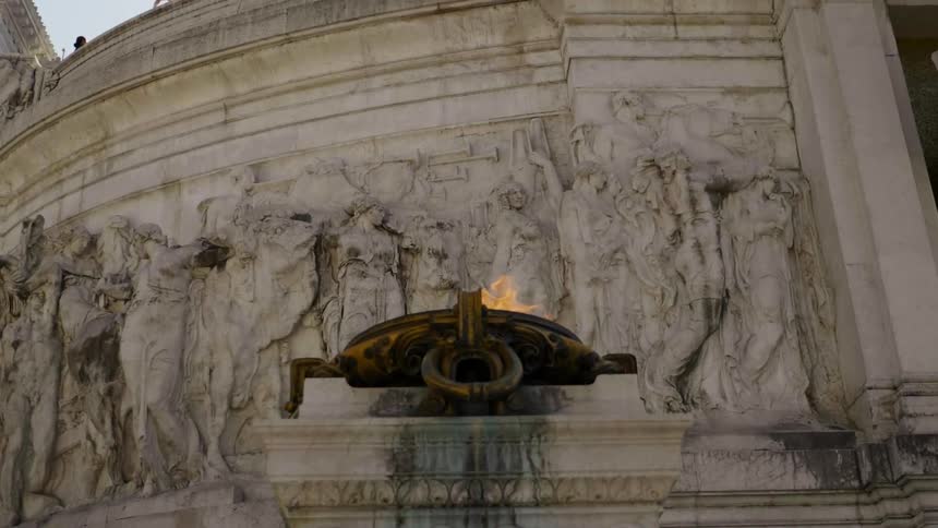 A fire burns in a cauldron on a column outside a building