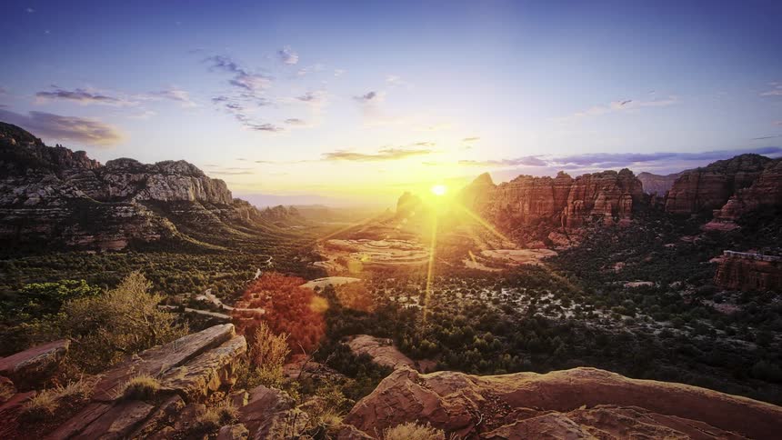 The sun sets and clouds pass by a wide desert landscape with rocky peaks in the distance