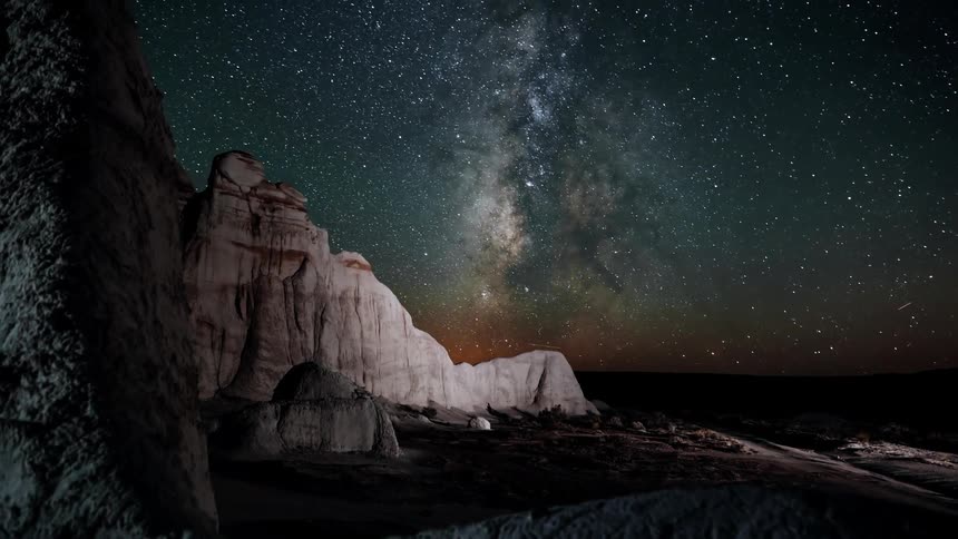 The starry night sky passes over rock formations in the desert