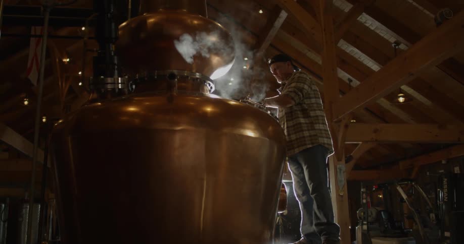 A distillery worker closes and opens equipment door as steam escapes