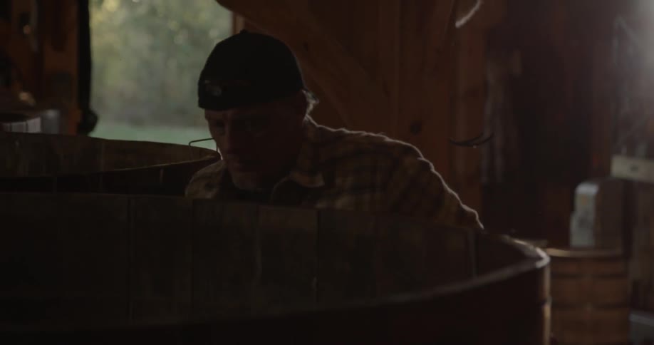 A distillery worker approaches a large vat with a graduated cylinder in hand