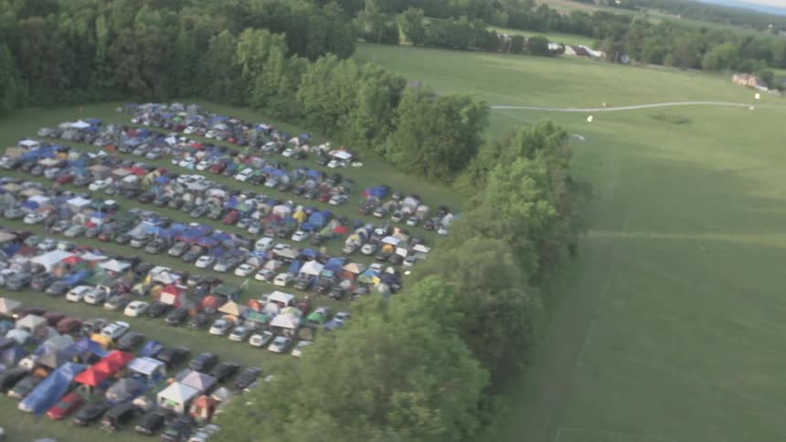 Dozens of vehicles are parked on a lawn surrounded by trees