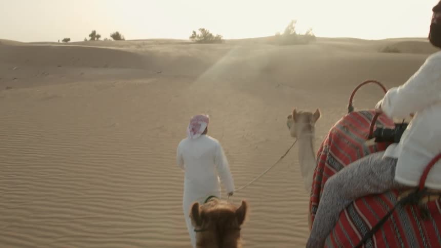 One person rides a camel and another leads a camel through the desert