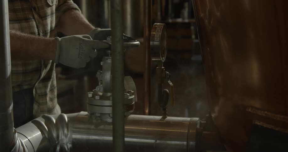 A distillery worker stands near steamy equipment and turns a wheel