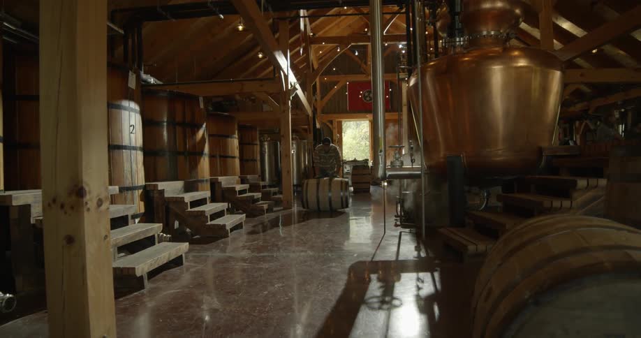 Steam rising from pipes in a brewery as a man rolls a barrel down the aisle