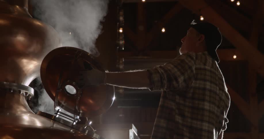 A man stands near steamy distillery equipment