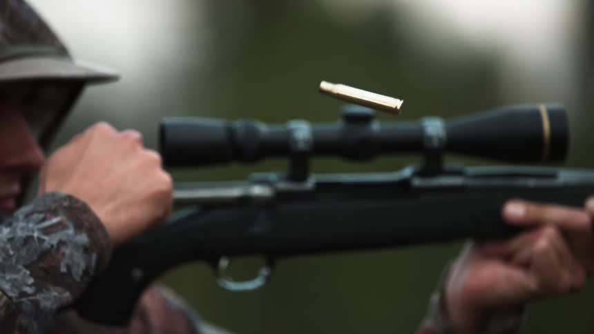 A man in camouflage fires a rifle and a bullet casing flies through the air