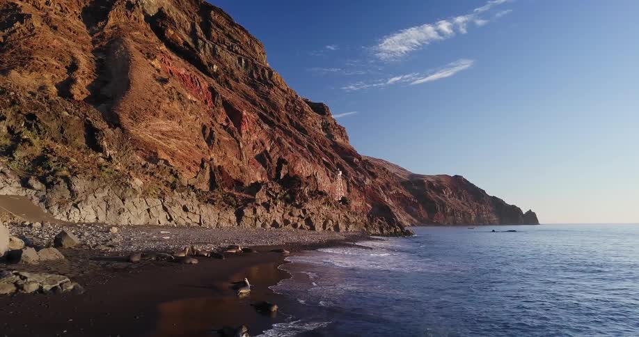 Ocean water is rolling in and breaking along the shoreline beneath a rocky mountain. of a rocky mountain