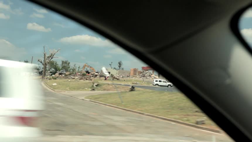A car travelling past damage done by a tornado with a still standing cross in the rubble