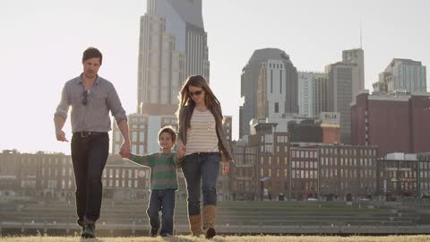A man, woman and child are holding hands walking through a field with a cityscape in the background