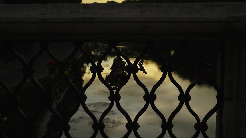 A cloudy, dusk sky is reflecting in a river beyond a bridge fence with locks on it