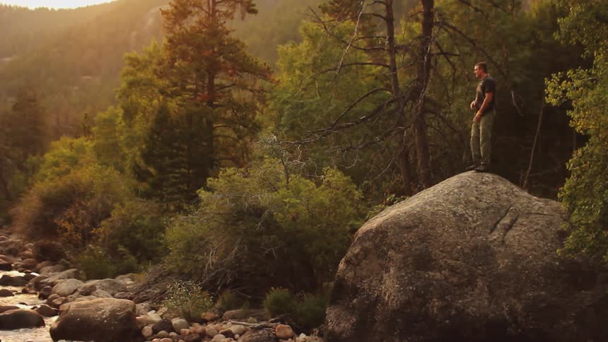 Man stands on a large rock overlooking a small stream in the woods