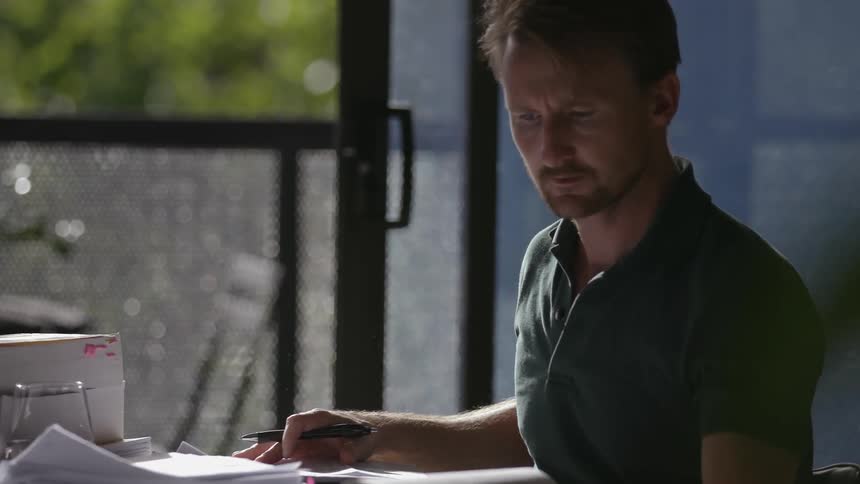 A man flips through a book while sitting at a small table with stacks of papers