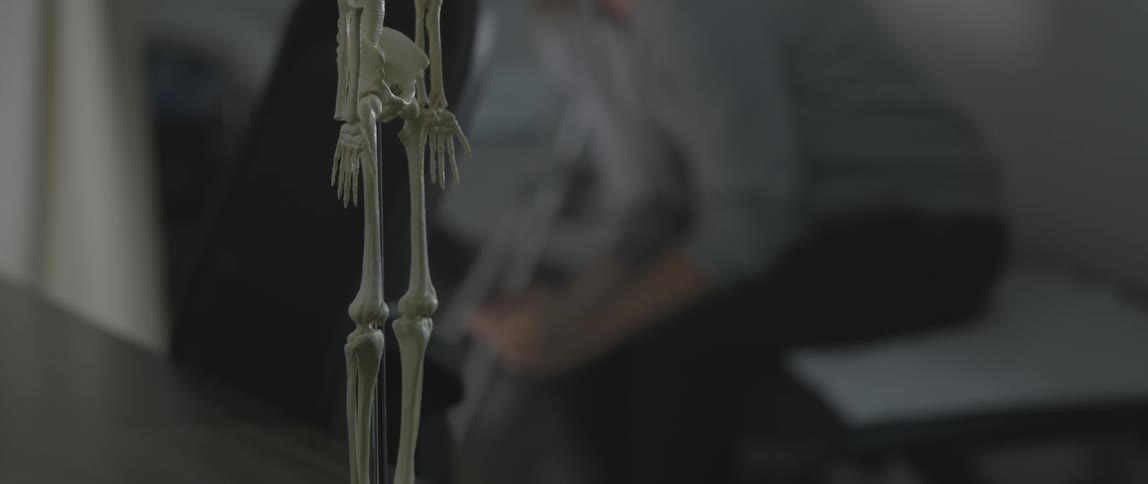 A woman sits wringing her hands in a room with a skeleton