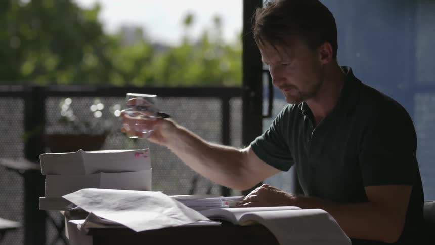 Man writes on a notebook looking out of a book and takes a sip of water