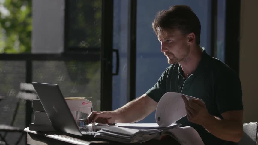 A man types on a laptop with a stack of papers on the small table