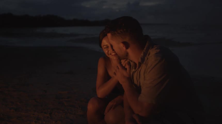 A man and woman squat down near a campfire on the beach