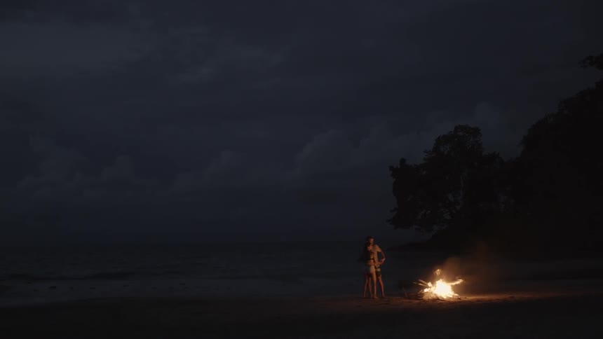 A couple stands on a beach near a fire during a storm