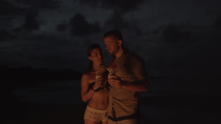 A man and woman drink while standing on a beach at night