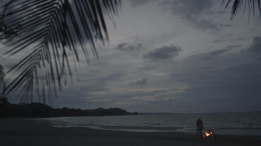 Two people stand beside a bonfire on a beach