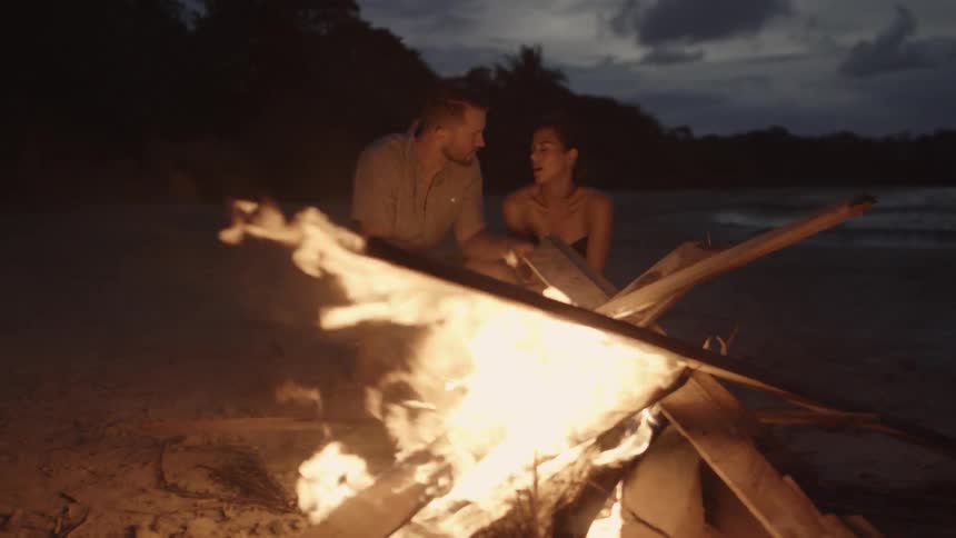 A man and woman talk by the campfire on the shore