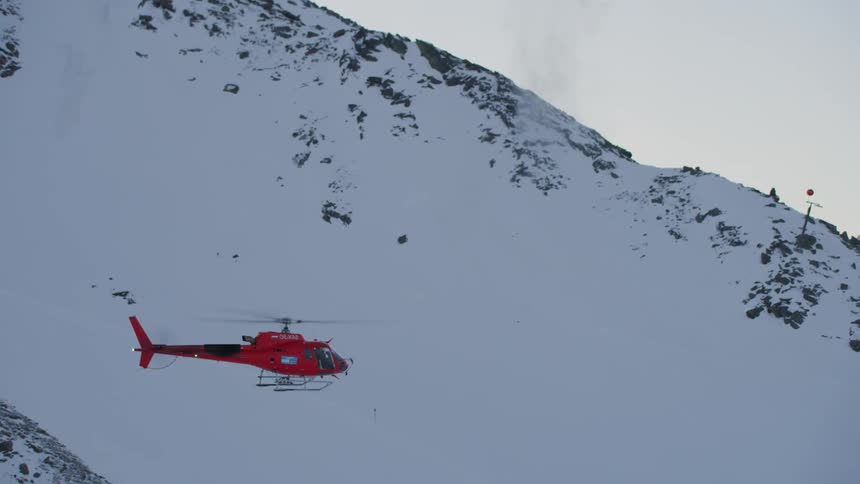 A helicopter hovers near a snow-covered mountainside as a landslide charge is detonated