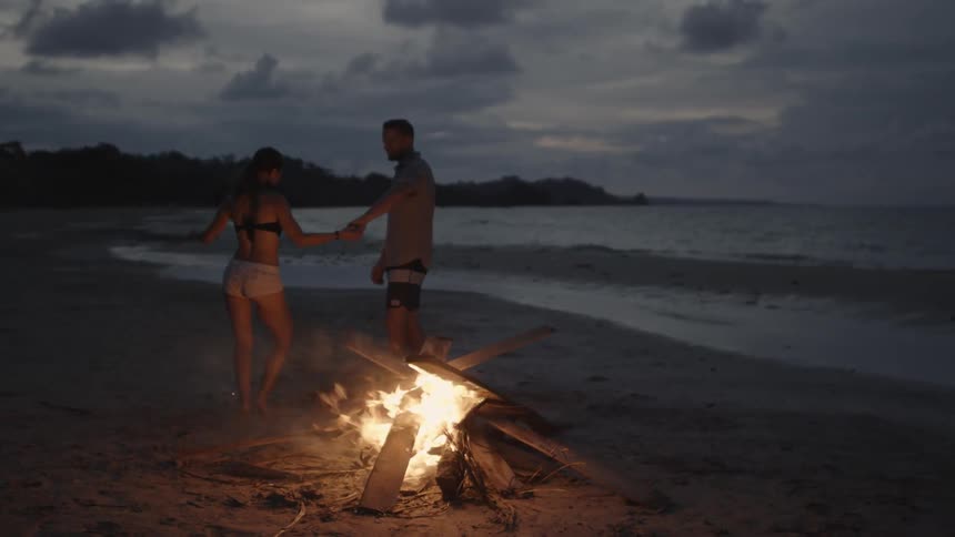 A man and woman dance beside a beach bonfire