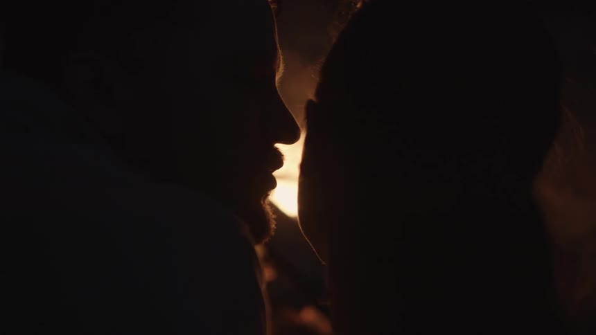 A man kisses a woman's cheek while they sit at a campfire