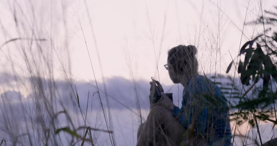 A woman sitting near tall grass eats from a bowl