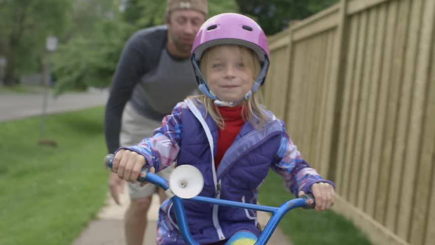A man helps a young girl ride a bicycle on a residential sidewalk