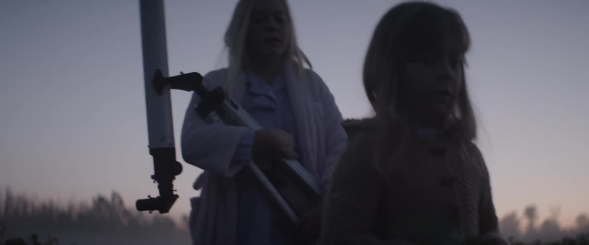 Two girls carry a telescope through a rural field at sunset