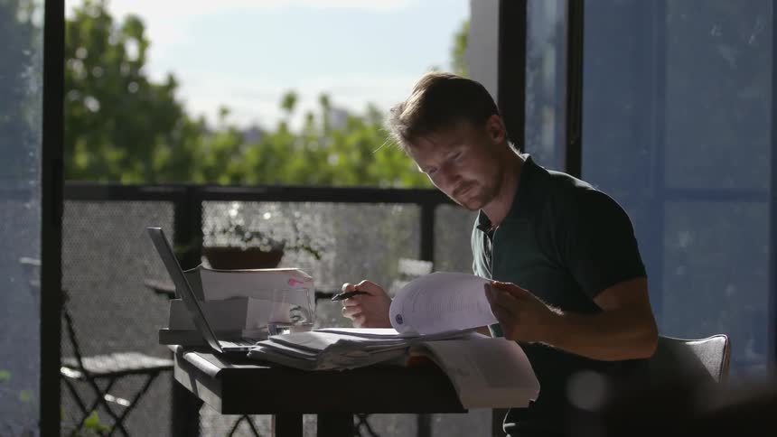 Man sits at a small desk with a laptop writing on papers