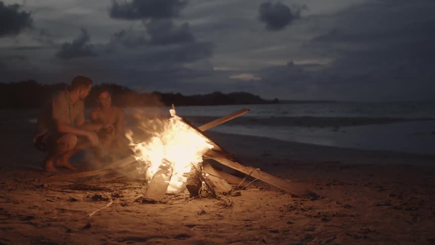 A man and woman sit next to a bonfire on the beach