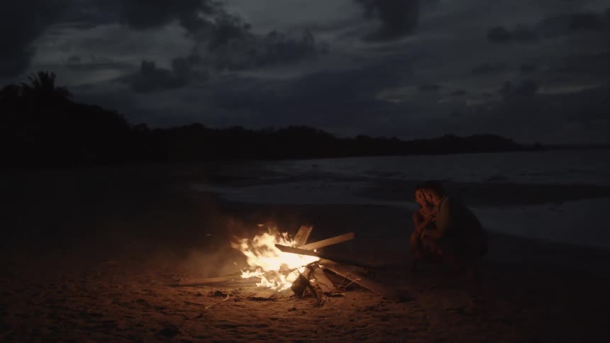 Man and woman sitting in front of a fire at the beach at night